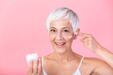 Beautiful mature woman holding jar of moisturizer and looking at camera. Happy senior woman holding a bottle of anti-aging lotion isolated over pink background. Anti aging and beauty treatment.