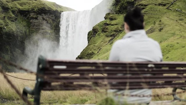 Woman looking at a waterfall, sitting on a bench, Skogafoss, Iceland - 4k