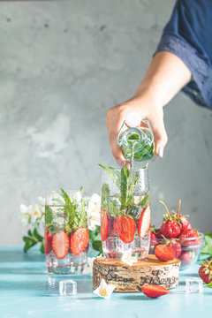 Women hand is pouring detox water from glass bottle to glass with strawberries, ice and mint. Summer fresh Mint soda cocktail, selective focus.