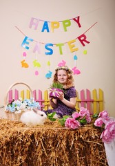 a child- a little girl in a dress with a flower wreath on her head smiles and holds a basket with greenery on the background of an Easter Bunny, hay and the inscription 