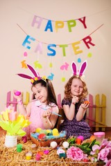 two little girl friends in fancy dresses and with Bunny ears on their heads smile, play and paint Easter eggs on the background of decor, hay, flowers, peonies and the inscription 