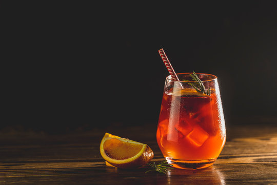 Glass with water drops of Italian aperol spritz cocktail with orange slices, ice and minton dark wooden table with amazing backlight. Milano spritzer alcoholic cocktail with red bitter