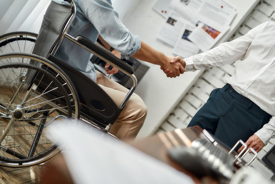 Cropped view of male office worker in a wheelchair shaking hand to his colleague while working together in the creative office. Disability concept