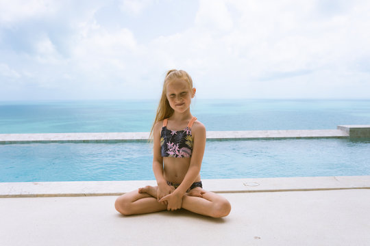 Little and beautiful girl sits by the pool in a bathing suit in lotus position. Morning yoga and calm. Sports practice and self-development. Healthy lifestyle