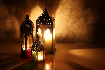 Ornamental Arabic lanterns with burning candles on table glowing at night. Festive greeting card, invitation for Muslim holy month Ramadan Kareem. Iftar dinner background with golden glow. - obrazy, fototapety, plakaty