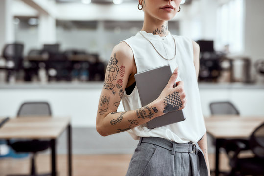 At work. Cropped photo of young stylish business woman with tattoos on her arm holding digital tablet while standing in the modern working space