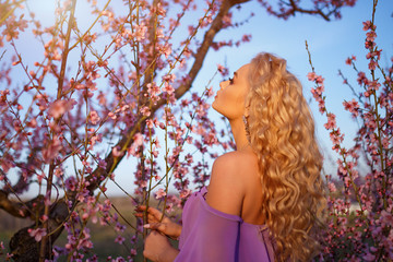 Beautiful young blonde woman with wavy hair posing with blooming peach trees against the sky