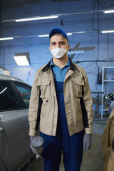 Vertical medium long shot portrait of professional car painter wearing protective workwear and mask on face looking at camera