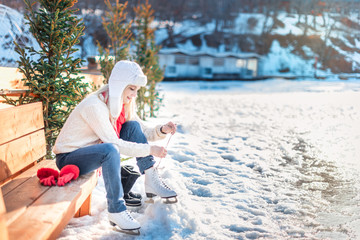 Young beautiful woman in white sweater sits on a bench and puts on figure skates early in the morning. Winter outdoor activities concept. Leisure and lifestyle. Background with copy space.