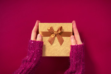 Female hands holding present with golden bow on pink background. Festive backdrop for holidays: Birthday, Valentines day, Christmas, New Year. Flat lay style. Banner