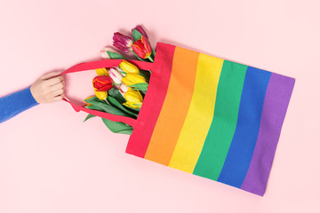 Female hand holding a LGBTQ pride rainbow flag canvas tote bag with tulip flowers on pink. top view banner or backdrop background. Homophobia, Transgender or lesbian visibility day and awareness