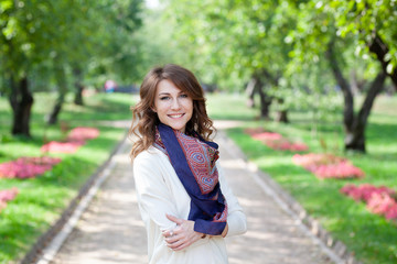 Obraz premium beautiful young brunette smiling happy woman in white jumper and with blue scarf in spring green park. trees and flowers are in background. girl's arms crossed on her chest in a closed position