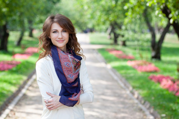 Obraz premium beautiful young brunette in white jumper and with blue scarf in spring green park. alley with trees and flowers are in background. girl's arms crossed on her chest in a closed position