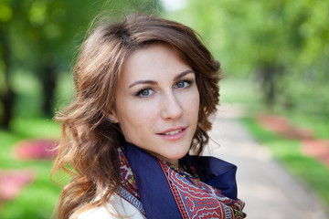 Fototapeta premium close-up portrait of beautiful young brunette woman in white jumper and with blue scarf in spring green park. alley with trees and flowers are in background.