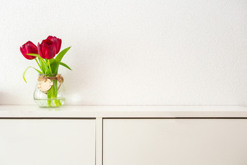Red tulips in tall glass jar on white table against white wall modern bright design, spring concept