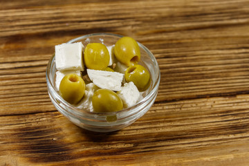 Feta cheese cubes with green olives in glass bowl on a wooden table