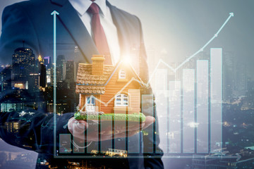 Double exposure of businessman holding house model in hand and animate of growing chart with city...