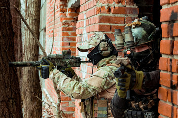 Two men in camouflage cloth and black uniform with machineguns stand beside brick wall. Soldiers...
