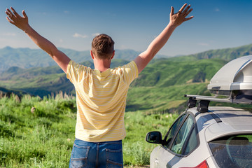 A road traveler stands next to a car and rejoices looking at the beautiful mountains