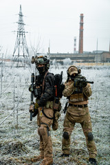 Fototapeta premium Men in camouflage cloth and black uniform with machineguns with factory on background