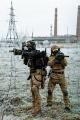 Obraz premium Men in camouflage cloth and black uniform with machineguns with factory on background. Soldiers with muchinegun aims aiming