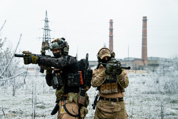 Men in camouflage cloth and black uniform with machineguns side to side with factory on background. Soldiers with muchinegun aims aiming