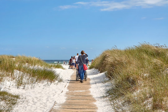 Dueodde, Bornholm island, Denmark - July 01, 2019. Woman pushing wheelchair on wooden boardwalk through sand dune towards Baltic Sea beach,