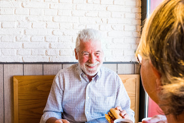 couple of two seniors eating and drinking in a restaurant of fast food together - mature man holding an hamburger and prepared to eat it looking at his wife