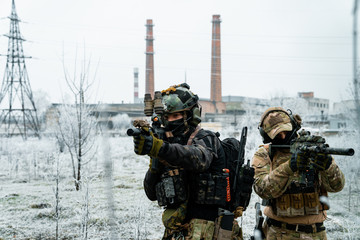 Men in camouflage cloth and black uniform with machineguns side to side with factory on background. Soldiers with muchinegun aims aiming