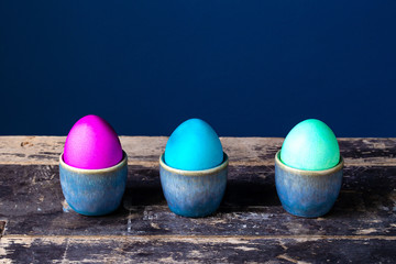Colorful bright Easter eggs hand painted in egg cups on a blue background on wooden shelf Holiday spring card