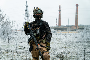 Man in black camouflage uniform hold machinegun. Soldier in the winter with factory on background. Horizontal photo.