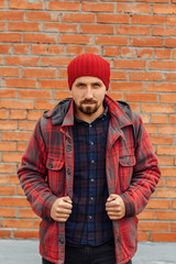 Young bearded man in red coat and hat. Guy with beard is standing in casual clothes against a brick wall and looking at camera.