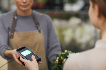 Cropped portrait of female customer paying via smartphone in flower shop, focus on smartphone screen, copy space