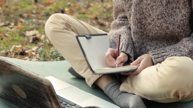 Female hiker sitting with crossed legs in front of laptop on camping mat in forest and taking notes about traveling impressions in notebook 