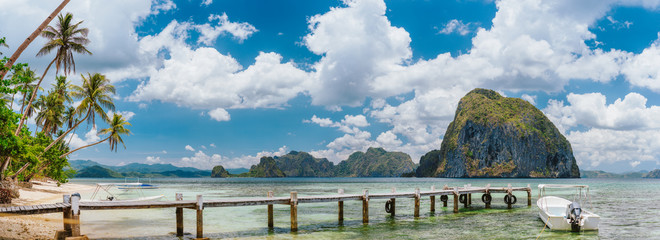 Beautiful tropical panorama of El Nido coastline, with jetty pier, palm trees, boat, huge limestone island and white clouds . Holiday and paradise island vacation concept
