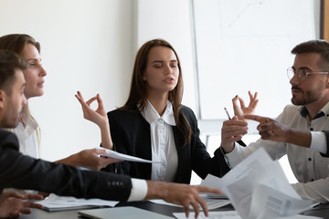 Mindful young business woman meditating, managing stress, ignoring annoyed arguing colleagues at workplace. Peaceful young female manager calming down during stressful working day at company office. © fizkes