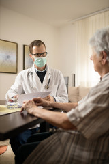 Serious young Caucasian doctor wearing safety mask consulting and advising senior white-haired woman during house call medical visit