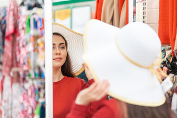 Fototapeta na wymiar A portrait of pretty Caucasian woman tries on a straw hat in front of a mirror in a store. Rear view, face reflected in the mirror. Shopping concept