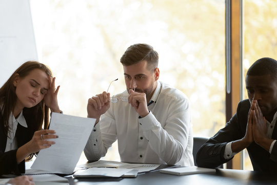 Mixed race frustrated group of employees feeling unmotivated fatigued. Young unhappy woman suffering from headache due to overload, millennial overwhelmed diverse businessmen distracted from job.
