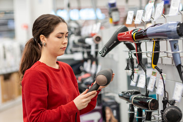 Caucasian young woman with long hair, holding a hair dryer. In the background, shelves with...