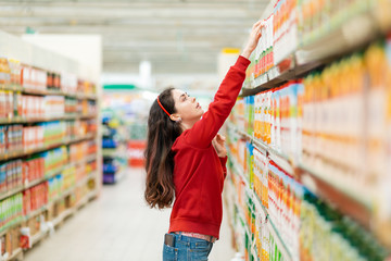 A young Caucasian woman in a red sweater reaches for a juice box on the top shelf. In the background, blurred rows of store shelves. The concept of buying products in a supermarket