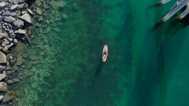 Paddle boarder goes along a rocky shoreline and dock over vibrant clear blue-green water.