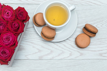 Red roses in gift box on white wooden table. Cup of tea and chocolate macaroons on a plate. Top view. Sweet dessert as a present on a table, minimal style.