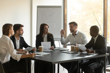 Concentrated serious mixed race young business employees team listening to confident male leader, explaining project details or problems, discussing reports during brainstorming meeting at office.