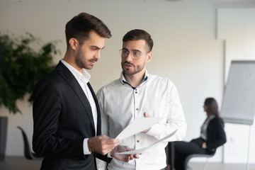 Two concentrated businessmen standing with paper documents, discussing project results, company marketing strategy at office. Young intern explaining work report to serious team leader at workplace.