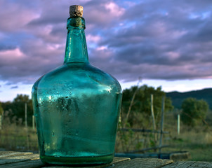 Fototapeta na wymiar Botella antigua de cristal en tonos verdeazulados con fondo de nubes azul violetas en el campo al aire libre
