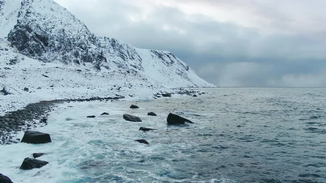 aerial view of snowy mountain slope overlooking sea drone flying low over water and rising up eggum beach lofoten