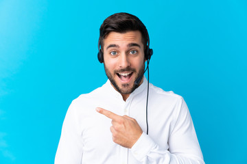 Telemarketer man working with a headset over isolated blue background pointing finger to the side