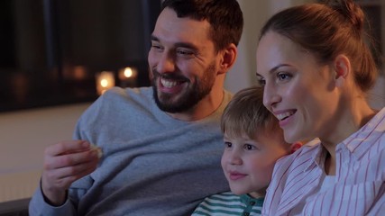 family, leisure and people concept - happy smiling father, mother and little son eating popcorn and watching tv at home in evening - Powered by Adobe