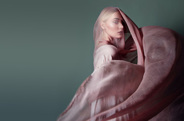 A beautiful young tender girl in a chiffon dress is covered with a drapery made of thin fabric that develops in the wind.